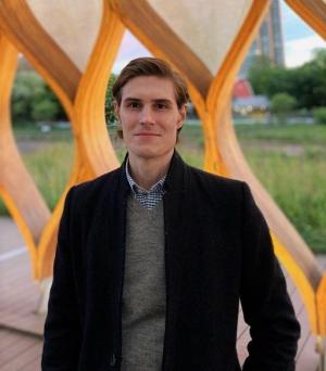 man in blazer and sweater standing in front of a geometric sculpture outside