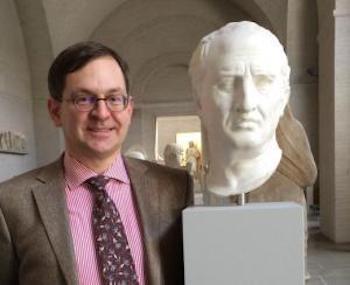 Man wearing round glasses and a suit and tie standing next to a Roman bust