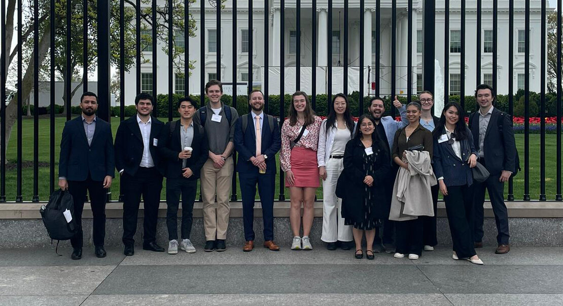 A group of students stand in front of a large black fence with the north face of the US White House in the background.