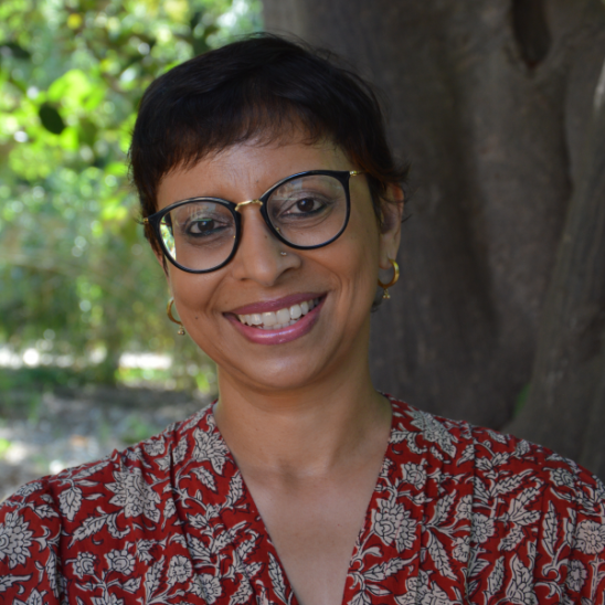headshot of a woman with short hair and glasses, wearing a red patterned top, standing in front of a tree